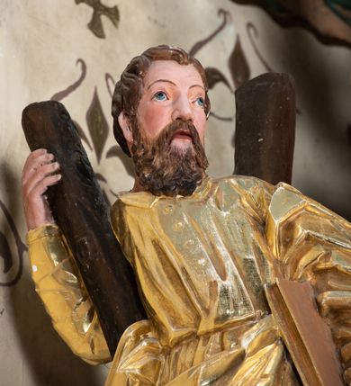 Zdjęcie nr 6: Rzeźba św. Andrzeja ścięta z tyłu i drążona, ustawiona na półkolistym i zielonym cokole. Święty ukazany jest frontalnie w delikatnym kontrapoście. W lewej dłoni trzyma, przytulając do biodra, księgę, a prawą obejmuje i opiera się o belkę krzyża w kształcie litery „X” stojącego za nim. Twarz pociągła z dużym nosem i szeroko rozstawionymi oczami, okolona długą i gęstą brodą oraz krótkimi i kręconymi włosami. Święty ubrany jest w suknię przewiązaną w talii z wąskimi rękawami i kołnierzykiem, zapiętą u góry na cztery guziki, a także płaszcz przewiązany przez lewe ramię i opadający na prawą stronę wzdłuż bioder; na stopach ma sandały. Tkaniny obficie drapowane w proste fałdy, płaszcz z wywiniętym na prawym boku brzegiem. Polichromia naturalistyczna w odsłoniętych partiach ciała, szaty złocone.