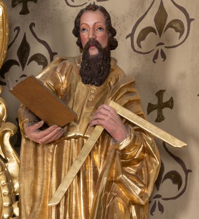 Zdjęcie nr 1: Rzeźba św. Tomasza ścięta z tyłu i drążona, ustawiona na półkolistym i zielonym cokole. Święty ukazany jest frontalnie, delikatnie pochylony w lewo. W prawej dłoni trzyma otwartą księgę, a W lewej ugiętej w łokciu węgielnicę. Twarz podłużna o dużym nosie i oczach, okolona długą i falistą brodą oraz półdługimi kręconymi włosami, zasłaniającymi uszy. Ubrany jest w długą suknię przewiązaną w talii z wąskimi rękawami i kołnierzykiem, zapiętą u góry na guziki, a także płaszcz zarzucony na ramiona i zawinięty wokół lewego przedramienia; stopy bose. Tkaniny są obficie drapowane w rurkowate i równoległe fałdy. Polichromia jest naturalistyczna w odsłoniętych partiach ciała, a szaty złocone.
