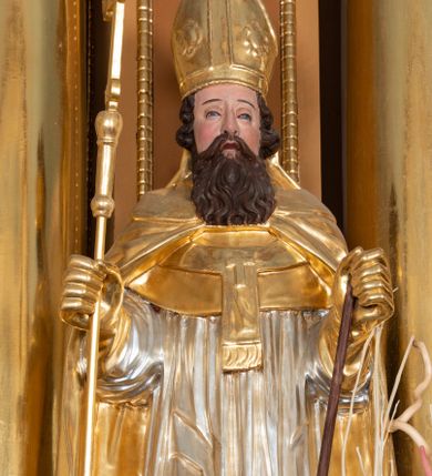 Zdjęcie nr 1: Rzeźba św. Wojciecha ścięta z tyłu i drążona, ustawiona na półkolistym i zielonym cokole. Święty ukazany jest frontalnie, w prawej dłoni trzyma krzyż, a w lewej wiosło. Twarz podłużna o dużym nosie i oczach, okolona długą i falistą brodą oraz półdługimi kręconymi włosami, zasłaniającymi uszy. Ubrany jest w srebrzoną albę i rokietę oraz w złocony paliusz, mucet, kapę, rękawice i infułę; na stopach ma założone czarne, pełne buty. Tkaniny są obficie drapowane w rurkowate i równoległe fałdy, u spodu łamane. Polichromia jest naturalistyczna w odsłoniętych partiach ciała.











