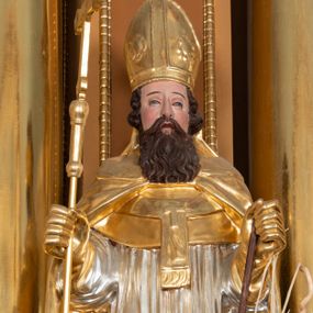 Zdjęcie nr 1: Rzeźba św. Wojciecha ścięta z tyłu i drążona, ustawiona na półkolistym i zielonym cokole. Święty ukazany jest frontalnie, w prawej dłoni trzyma krzyż, a w lewej wiosło. Twarz podłużna o dużym nosie i oczach, okolona długą i falistą brodą oraz półdługimi kręconymi włosami, zasłaniającymi uszy. Ubrany jest w srebrzoną albę i rokietę oraz w złocony paliusz, mucet, kapę, rękawice i infułę; na stopach ma założone czarne, pełne buty. Tkaniny są obficie drapowane w rurkowate i równoległe fałdy, u spodu łamane. Polichromia jest naturalistyczna w odsłoniętych partiach ciała.











