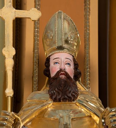 Zdjęcie nr 4: Rzeźba św. Wojciecha ścięta z tyłu i drążona, ustawiona na półkolistym i zielonym cokole. Święty ukazany jest frontalnie, w prawej dłoni trzyma krzyż, a w lewej wiosło. Twarz podłużna o dużym nosie i oczach, okolona długą i falistą brodą oraz półdługimi kręconymi włosami, zasłaniającymi uszy. Ubrany jest w srebrzoną albę i rokietę oraz w złocony paliusz, mucet, kapę, rękawice i infułę; na stopach ma założone czarne, pełne buty. Tkaniny są obficie drapowane w rurkowate i równoległe fałdy, u spodu łamane. Polichromia jest naturalistyczna w odsłoniętych partiach ciała.











