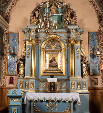 Zdjęcie nr 12: Obraz Matki Boskiej z Dzieciątkiem w kształcie stojącego prostokąta. Maria ukazana w półpostaci, delikatnie zwrócona w lewo z Dzieciątkiem Jezus na lewym ręku, z głową przytuloną do jego twarzy, prawą dłonią trzymająca jego lewą rączkę. Twarz Marii pełna, nos długi, duże i brązowe oczy oraz drobne usta. Ubrana jest w niebieską suknię z długimi rękawami, ozdobioną na brzegu złoconą bordiurą oraz ciemnoczerwony maforion , malowanym światłocieniowo z zieloną podszewką i z lewą połą zarzuconą na prawe ramię. Płaszcz ozdobiony jest trzema gwiazdami: na czole, na dekolcie po lewej stronie i na prawym ramieniu; a na brzegach dekorowany jest ozdobną lamówką z filigranowymi frędzlami. Dzieciątko siedzi na ramieniu Matki, zwrócone trzy czwarte w prawo, ma skrzyżowane nóżki, a lewą rękę trzyma w prawej dłoni Matki Boskiej. Ubrane jest w srebrną sukienkę z długimi i szerokimi rękawami, przewiązaną w talii jasnobeżową tkaniną oraz złoconą szatę wierzchnią, bez rękawów, skrzyżowaną na piersi, ozdobioną szrafowaniem. Twarz Jezusa pełna o pucułowatych policzkach i podkreślonych podbródku z długim nosem, małymi ustami i dużymi ciemnymi oczami. Karnacje obu postaci ciemne z zarumienionymi policzkami. Na głowach mają założone pełne korony zdobione fioletowymi, niebieskimi i czerwonymi kamieniami; a wokół głów koliste i złocone nimby. Na wysokości głowy Matki Bożej, po lewej stronie znajdują się tłoczone abrewiacje: „MP ΘY”, a po prawej: „IX XC”.  Ornament tła wyciskany w gruncie, złocony o układzie kandelabrowym, roślinnym z motyw palmetowym. Na odwrociu napis: „obraz podług trad(ycji) z IX w(wieku) konserw(owany) z XIV w(ieku), odnow(iony) i zakonserw(owany) 1963 kons(erwator) Zawadzki T(adeusz) Dzierża z Kr(akowa) Prob(oszcz) X(iądz) J(ózef) Długopolski kość. Mika Fr.” Wizerunek umieszczony jest w złoconej, profilowanej ramie, od wewnątrz zdobionej pasem kimationu.

