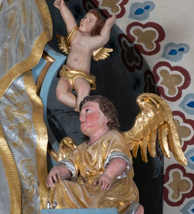 Zdjęcie nr 11: Zwieńczenie ołtarza o fantazyjnym kształcie zbliżonym do stojącego prostokąta z płaskorzeźbioną sceną ofiarowania szkaplerza św. Szymonowi Stockowi. U góry zwieńczenia podpięta srebrzona kotara o złoconym brzegu, spływająca na jego boki. W górnej partii, po bokach para aniołków z wzniesionymi do góry rękami oraz dwie uskrzydlone główki anielskie. Na szczycie nastawy zatknięta jest promienista gloria złożona z wiązek promieni o nierównej długości z monogramem maryjnym otoczonym przez wieniec z obłoków. Pole zwieńczenia dwustrefowe, oddzielone pasem obłoków. W dolnej strefie, po lewej stronie rzeźba św. Szymona Stocka. Święty ukazany jest w pozycji klęczącej z prawego profilu z wniesionymi na wysokości piersi rękami i głową skierowaną ku Matce Bożej. Twarz podłużna z długim nosem i silnie zarumienionymi policzkami. Głowę okala długa i siwa broda oraz tonsura. Święty ubrany jest w brązowy habit i płaszcz karmelitański. Po prawej stronie obrazu płaskorzeźbione, zielone wzniesienia i duże drzewo. W tle malowany pejzaż miejski i jasnobłękitne niebo. W górnej strefie siedząca na obłokach Matka Boska z Dzieciątkiem. W prawej dłoni trzyma szkaplerze zdobione greckim krzyżykiem oraz gorejącym sercem, przebitym mieczem, a lewą ręką przytrzymuje siedzące na jej kolanach Dzieciątko Jezus. Maria ma podłużną twarz z niewielkimi ustami, długim nosem i jasnoniebieskimi oczami, okoloną ciemnobrązowymi włosami, opadającymi na plecy. Ubrana jest w złocone szaty: suknię i płaszcz, obficie drapowane. Na głowie ma założony srebrzony welon oraz pełną koronę, a wokół niej wieniec z 12 gwiazd. Polichromia struktury zwieńczenia w kolorze jasnoniebieskim, dekoracje ornamentalne oraz detale złocone i srebrzone. Polichromia w odsłoniętych partiach ciała figur naturalistyczna.

