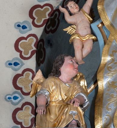 Zdjęcie nr 10: Zwieńczenie ołtarza o fantazyjnym kształcie zbliżonym do stojącego prostokąta z płaskorzeźbioną sceną ofiarowania szkaplerza św. Szymonowi Stockowi. U góry zwieńczenia podpięta srebrzona kotara o złoconym brzegu, spływająca na jego boki. W górnej partii, po bokach para aniołków z wzniesionymi do góry rękami oraz dwie uskrzydlone główki anielskie. Na szczycie nastawy zatknięta jest promienista gloria złożona z wiązek promieni o nierównej długości z monogramem maryjnym otoczonym przez wieniec z obłoków. Pole zwieńczenia dwustrefowe, oddzielone pasem obłoków. W dolnej strefie, po lewej stronie rzeźba św. Szymona Stocka. Święty ukazany jest w pozycji klęczącej z prawego profilu z wniesionymi na wysokości piersi rękami i głową skierowaną ku Matce Bożej. Twarz podłużna z długim nosem i silnie zarumienionymi policzkami. Głowę okala długa i siwa broda oraz tonsura. Święty ubrany jest w brązowy habit i płaszcz karmelitański. Po prawej stronie obrazu płaskorzeźbione, zielone wzniesienia i duże drzewo. W tle malowany pejzaż miejski i jasnobłękitne niebo. W górnej strefie siedząca na obłokach Matka Boska z Dzieciątkiem. W prawej dłoni trzyma szkaplerze zdobione greckim krzyżykiem oraz gorejącym sercem, przebitym mieczem, a lewą ręką przytrzymuje siedzące na jej kolanach Dzieciątko Jezus. Maria ma podłużną twarz z niewielkimi ustami, długim nosem i jasnoniebieskimi oczami, okoloną ciemnobrązowymi włosami, opadającymi na plecy. Ubrana jest w złocone szaty: suknię i płaszcz, obficie drapowane. Na głowie ma założony srebrzony welon oraz pełną koronę, a wokół niej wieniec z 12 gwiazd. Polichromia struktury zwieńczenia w kolorze jasnoniebieskim, dekoracje ornamentalne oraz detale złocone i srebrzone. Polichromia w odsłoniętych partiach ciała figur naturalistyczna.

