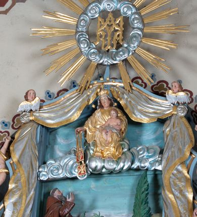 Zdjęcie nr 7: Zwieńczenie ołtarza o fantazyjnym kształcie zbliżonym do stojącego prostokąta z płaskorzeźbioną sceną ofiarowania szkaplerza św. Szymonowi Stockowi. U góry zwieńczenia podpięta srebrzona kotara o złoconym brzegu, spływająca na jego boki. W górnej partii, po bokach para aniołków z wzniesionymi do góry rękami oraz dwie uskrzydlone główki anielskie. Na szczycie nastawy zatknięta jest promienista gloria złożona z wiązek promieni o nierównej długości z monogramem maryjnym otoczonym przez wieniec z obłoków. Pole zwieńczenia dwustrefowe, oddzielone pasem obłoków. W dolnej strefie, po lewej stronie rzeźba św. Szymona Stocka. Święty ukazany jest w pozycji klęczącej z prawego profilu z wniesionymi na wysokości piersi rękami i głową skierowaną ku Matce Bożej. Twarz podłużna z długim nosem i silnie zarumienionymi policzkami. Głowę okala długa i siwa broda oraz tonsura. Święty ubrany jest w brązowy habit i płaszcz karmelitański. Po prawej stronie obrazu płaskorzeźbione, zielone wzniesienia i duże drzewo. W tle malowany pejzaż miejski i jasnobłękitne niebo. W górnej strefie siedząca na obłokach Matka Boska z Dzieciątkiem. W prawej dłoni trzyma szkaplerze zdobione greckim krzyżykiem oraz gorejącym sercem, przebitym mieczem, a lewą ręką przytrzymuje siedzące na jej kolanach Dzieciątko Jezus. Maria ma podłużną twarz z niewielkimi ustami, długim nosem i jasnoniebieskimi oczami, okoloną ciemnobrązowymi włosami, opadającymi na plecy. Ubrana jest w złocone szaty: suknię i płaszcz, obficie drapowane. Na głowie ma założony srebrzony welon oraz pełną koronę, a wokół niej wieniec z 12 gwiazd. Polichromia struktury zwieńczenia w kolorze jasnoniebieskim, dekoracje ornamentalne oraz detale złocone i srebrzone. Polichromia w odsłoniętych partiach ciała figur naturalistyczna.

