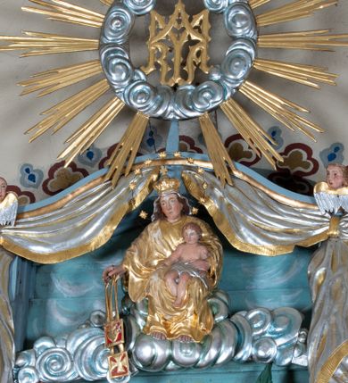 Zdjęcie nr 1: Zwieńczenie ołtarza o fantazyjnym kształcie zbliżonym do stojącego prostokąta z płaskorzeźbioną sceną ofiarowania szkaplerza św. Szymonowi Stockowi. U góry zwieńczenia podpięta srebrzona kotara o złoconym brzegu, spływająca na jego boki. W górnej partii, po bokach para aniołków z wzniesionymi do góry rękami oraz dwie uskrzydlone główki anielskie. Na szczycie nastawy zatknięta jest promienista gloria złożona z wiązek promieni o nierównej długości z monogramem maryjnym otoczonym przez wieniec z obłoków. Pole zwieńczenia dwustrefowe, oddzielone pasem obłoków. W dolnej strefie, po lewej stronie rzeźba św. Szymona Stocka. Święty ukazany jest w pozycji klęczącej z prawego profilu z wniesionymi na wysokości piersi rękami i głową skierowaną ku Matce Bożej. Twarz podłużna z długim nosem i silnie zarumienionymi policzkami. Głowę okala długa i siwa broda oraz tonsura. Święty ubrany jest w brązowy habit i płaszcz karmelitański. Po prawej stronie obrazu płaskorzeźbione, zielone wzniesienia i duże drzewo. W tle malowany pejzaż miejski i jasnobłękitne niebo. W górnej strefie siedząca na obłokach Matka Boska z Dzieciątkiem. W prawej dłoni trzyma szkaplerze zdobione greckim krzyżykiem oraz gorejącym sercem, przebitym mieczem, a lewą ręką przytrzymuje siedzące na jej kolanach Dzieciątko Jezus. Maria ma podłużną twarz z niewielkimi ustami, długim nosem i jasnoniebieskimi oczami, okoloną ciemnobrązowymi włosami, opadającymi na plecy. Ubrana jest w złocone szaty: suknię i płaszcz, obficie drapowane. Na głowie ma założony srebrzony welon oraz pełną koronę, a wokół niej wieniec z 12 gwiazd. Polichromia struktury zwieńczenia w kolorze jasnoniebieskim, dekoracje ornamentalne oraz detale złocone i srebrzone. Polichromia w odsłoniętych partiach ciała figur naturalistyczna.

