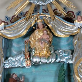 Zdjęcie nr 1: Zwieńczenie ołtarza o fantazyjnym kształcie zbliżonym do stojącego prostokąta z płaskorzeźbioną sceną ofiarowania szkaplerza św. Szymonowi Stockowi. U góry zwieńczenia podpięta srebrzona kotara o złoconym brzegu, spływająca na jego boki. W górnej partii, po bokach para aniołków z wzniesionymi do góry rękami oraz dwie uskrzydlone główki anielskie. Na szczycie nastawy zatknięta jest promienista gloria złożona z wiązek promieni o nierównej długości z monogramem maryjnym otoczonym przez wieniec z obłoków. Pole zwieńczenia dwustrefowe, oddzielone pasem obłoków. W dolnej strefie, po lewej stronie rzeźba św. Szymona Stocka. Święty ukazany jest w pozycji klęczącej z prawego profilu z wniesionymi na wysokości piersi rękami i głową skierowaną ku Matce Bożej. Twarz podłużna z długim nosem i silnie zarumienionymi policzkami. Głowę okala długa i siwa broda oraz tonsura. Święty ubrany jest w brązowy habit i płaszcz karmelitański. Po prawej stronie obrazu płaskorzeźbione, zielone wzniesienia i duże drzewo. W tle malowany pejzaż miejski i jasnobłękitne niebo. W górnej strefie siedząca na obłokach Matka Boska z Dzieciątkiem. W prawej dłoni trzyma szkaplerze zdobione greckim krzyżykiem oraz gorejącym sercem, przebitym mieczem, a lewą ręką przytrzymuje siedzące na jej kolanach Dzieciątko Jezus. Maria ma podłużną twarz z niewielkimi ustami, długim nosem i jasnoniebieskimi oczami, okoloną ciemnobrązowymi włosami, opadającymi na plecy. Ubrana jest w złocone szaty: suknię i płaszcz, obficie drapowane. Na głowie ma założony srebrzony welon oraz pełną koronę, a wokół niej wieniec z 12 gwiazd. Polichromia struktury zwieńczenia w kolorze jasnoniebieskim, dekoracje ornamentalne oraz detale złocone i srebrzone. Polichromia w odsłoniętych partiach ciała figur naturalistyczna.

