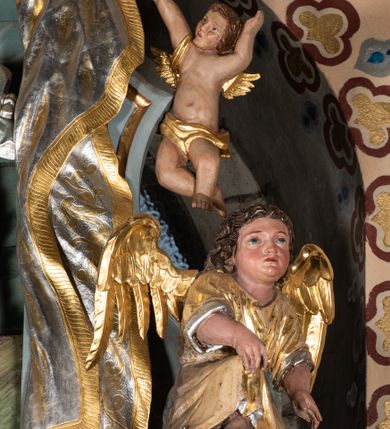 Zdjęcie nr 12: Zwieńczenie ołtarza o fantazyjnym kształcie zbliżonym do stojącego prostokąta z płaskorzeźbioną sceną ofiarowania szkaplerza św. Szymonowi Stockowi. U góry zwieńczenia podpięta srebrzona kotara o złoconym brzegu, spływająca na jego boki. W górnej partii, po bokach para aniołków z wzniesionymi do góry rękami oraz dwie uskrzydlone główki anielskie. Na szczycie nastawy zatknięta jest promienista gloria złożona z wiązek promieni o nierównej długości z monogramem maryjnym otoczonym przez wieniec z obłoków. Pole zwieńczenia dwustrefowe, oddzielone pasem obłoków. W dolnej strefie, po lewej stronie rzeźba św. Szymona Stocka. Święty ukazany jest w pozycji klęczącej z prawego profilu z wniesionymi na wysokości piersi rękami i głową skierowaną ku Matce Bożej. Twarz podłużna z długim nosem i silnie zarumienionymi policzkami. Głowę okala długa i siwa broda oraz tonsura. Święty ubrany jest w brązowy habit i płaszcz karmelitański. Po prawej stronie obrazu płaskorzeźbione, zielone wzniesienia i duże drzewo. W tle malowany pejzaż miejski i jasnobłękitne niebo. W górnej strefie siedząca na obłokach Matka Boska z Dzieciątkiem. W prawej dłoni trzyma szkaplerze zdobione greckim krzyżykiem oraz gorejącym sercem, przebitym mieczem, a lewą ręką przytrzymuje siedzące na jej kolanach Dzieciątko Jezus. Maria ma podłużną twarz z niewielkimi ustami, długim nosem i jasnoniebieskimi oczami, okoloną ciemnobrązowymi włosami, opadającymi na plecy. Ubrana jest w złocone szaty: suknię i płaszcz, obficie drapowane. Na głowie ma założony srebrzony welon oraz pełną koronę, a wokół niej wieniec z 12 gwiazd. Polichromia struktury zwieńczenia w kolorze jasnoniebieskim, dekoracje ornamentalne oraz detale złocone i srebrzone. Polichromia w odsłoniętych partiach ciała figur naturalistyczna.

