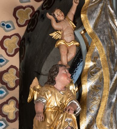 Zdjęcie nr 2: Zwieńczenie ołtarza o fantazyjnym kształcie zbliżonym do stojącego prostokąta z płaskorzeźbioną sceną ofiarowania szkaplerza św. Szymonowi Stockowi. U góry zwieńczenia podpięta srebrzona kotara o złoconym brzegu, spływająca na jego boki. W górnej partii, po bokach para aniołków z wzniesionymi do góry rękami oraz dwie uskrzydlone główki anielskie. Na szczycie nastawy zatknięta jest promienista gloria złożona z wiązek promieni o nierównej długości z monogramem maryjnym otoczonym przez wieniec z obłoków. Pole zwieńczenia dwustrefowe, oddzielone pasem obłoków. W dolnej strefie, po lewej stronie rzeźba św. Szymona Stocka. Święty ukazany jest w pozycji klęczącej z prawego profilu z wniesionymi na wysokości piersi rękami i głową skierowaną ku Matce Bożej. Twarz podłużna z długim nosem i silnie zarumienionymi policzkami. Głowę okala długa i siwa broda oraz tonsura. Święty ubrany jest w brązowy habit i płaszcz karmelitański. Po prawej stronie obrazu płaskorzeźbione, zielone wzniesienia i duże drzewo. W tle malowany pejzaż miejski i jasnobłękitne niebo. W górnej strefie siedząca na obłokach Matka Boska z Dzieciątkiem. W prawej dłoni trzyma szkaplerze zdobione greckim krzyżykiem oraz gorejącym sercem, przebitym mieczem, a lewą ręką przytrzymuje siedzące na jej kolanach Dzieciątko Jezus. Maria ma podłużną twarz z niewielkimi ustami, długim nosem i jasnoniebieskimi oczami, okoloną ciemnobrązowymi włosami, opadającymi na plecy. Ubrana jest w złocone szaty: suknię i płaszcz, obficie drapowane. Na głowie ma założony srebrzony welon oraz pełną koronę, a wokół niej wieniec z 12 gwiazd. Polichromia struktury zwieńczenia w kolorze jasnoniebieskim, dekoracje ornamentalne oraz detale złocone i srebrzone. Polichromia w odsłoniętych partiach ciała figur naturalistyczna.

