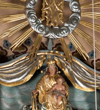 Zdjęcie nr 6: Zwieńczenie ołtarza o fantazyjnym kształcie zbliżonym do stojącego prostokąta z płaskorzeźbioną sceną ofiarowania szkaplerza św. Szymonowi Stockowi. U góry zwieńczenia podpięta srebrzona kotara o złoconym brzegu, spływająca na jego boki. W górnej partii, po bokach para aniołków z wzniesionymi do góry rękami oraz dwie uskrzydlone główki anielskie. Na szczycie nastawy zatknięta jest promienista gloria złożona z wiązek promieni o nierównej długości z monogramem maryjnym otoczonym przez wieniec z obłoków. Pole zwieńczenia dwustrefowe, oddzielone pasem obłoków. W dolnej strefie, po lewej stronie rzeźba św. Szymona Stocka. Święty ukazany jest w pozycji klęczącej z prawego profilu z wniesionymi na wysokości piersi rękami i głową skierowaną ku Matce Bożej. Twarz podłużna z długim nosem i silnie zarumienionymi policzkami. Głowę okala długa i siwa broda oraz tonsura. Święty ubrany jest w brązowy habit i płaszcz karmelitański. Po prawej stronie obrazu płaskorzeźbione, zielone wzniesienia i duże drzewo. W tle malowany pejzaż miejski i jasnobłękitne niebo. W górnej strefie siedząca na obłokach Matka Boska z Dzieciątkiem. W prawej dłoni trzyma szkaplerze zdobione greckim krzyżykiem oraz gorejącym sercem, przebitym mieczem, a lewą ręką przytrzymuje siedzące na jej kolanach Dzieciątko Jezus. Maria ma podłużną twarz z niewielkimi ustami, długim nosem i jasnoniebieskimi oczami, okoloną ciemnobrązowymi włosami, opadającymi na plecy. Ubrana jest w złocone szaty: suknię i płaszcz, obficie drapowane. Na głowie ma założony srebrzony welon oraz pełną koronę, a wokół niej wieniec z 12 gwiazd. Polichromia struktury zwieńczenia w kolorze jasnoniebieskim, dekoracje ornamentalne oraz detale złocone i srebrzone. Polichromia w odsłoniętych partiach ciała figur naturalistyczna.

