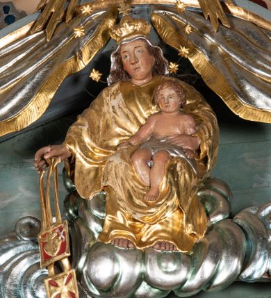 Zdjęcie nr 3: Zwieńczenie ołtarza o fantazyjnym kształcie zbliżonym do stojącego prostokąta z płaskorzeźbioną sceną ofiarowania szkaplerza św. Szymonowi Stockowi. U góry zwieńczenia podpięta srebrzona kotara o złoconym brzegu, spływająca na jego boki. W górnej partii, po bokach para aniołków z wzniesionymi do góry rękami oraz dwie uskrzydlone główki anielskie. Na szczycie nastawy zatknięta jest promienista gloria złożona z wiązek promieni o nierównej długości z monogramem maryjnym otoczonym przez wieniec z obłoków. Pole zwieńczenia dwustrefowe, oddzielone pasem obłoków. W dolnej strefie, po lewej stronie rzeźba św. Szymona Stocka. Święty ukazany jest w pozycji klęczącej z prawego profilu z wniesionymi na wysokości piersi rękami i głową skierowaną ku Matce Bożej. Twarz podłużna z długim nosem i silnie zarumienionymi policzkami. Głowę okala długa i siwa broda oraz tonsura. Święty ubrany jest w brązowy habit i płaszcz karmelitański. Po prawej stronie obrazu płaskorzeźbione, zielone wzniesienia i duże drzewo. W tle malowany pejzaż miejski i jasnobłękitne niebo. W górnej strefie siedząca na obłokach Matka Boska z Dzieciątkiem. W prawej dłoni trzyma szkaplerze zdobione greckim krzyżykiem oraz gorejącym sercem, przebitym mieczem, a lewą ręką przytrzymuje siedzące na jej kolanach Dzieciątko Jezus. Maria ma podłużną twarz z niewielkimi ustami, długim nosem i jasnoniebieskimi oczami, okoloną ciemnobrązowymi włosami, opadającymi na plecy. Ubrana jest w złocone szaty: suknię i płaszcz, obficie drapowane. Na głowie ma założony srebrzony welon oraz pełną koronę, a wokół niej wieniec z 12 gwiazd. Polichromia struktury zwieńczenia w kolorze jasnoniebieskim, dekoracje ornamentalne oraz detale złocone i srebrzone. Polichromia w odsłoniętych partiach ciała figur naturalistyczna.

