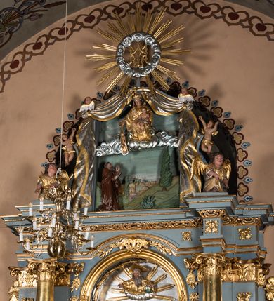 Zdjęcie nr 9: Zwieńczenie ołtarza o fantazyjnym kształcie zbliżonym do stojącego prostokąta z płaskorzeźbioną sceną ofiarowania szkaplerza św. Szymonowi Stockowi. U góry zwieńczenia podpięta srebrzona kotara o złoconym brzegu, spływająca na jego boki. W górnej partii, po bokach para aniołków z wzniesionymi do góry rękami oraz dwie uskrzydlone główki anielskie. Na szczycie nastawy zatknięta jest promienista gloria złożona z wiązek promieni o nierównej długości z monogramem maryjnym otoczonym przez wieniec z obłoków. Pole zwieńczenia dwustrefowe, oddzielone pasem obłoków. W dolnej strefie, po lewej stronie rzeźba św. Szymona Stocka. Święty ukazany jest w pozycji klęczącej z prawego profilu z wniesionymi na wysokości piersi rękami i głową skierowaną ku Matce Bożej. Twarz podłużna z długim nosem i silnie zarumienionymi policzkami. Głowę okala długa i siwa broda oraz tonsura. Święty ubrany jest w brązowy habit i płaszcz karmelitański. Po prawej stronie obrazu płaskorzeźbione, zielone wzniesienia i duże drzewo. W tle malowany pejzaż miejski i jasnobłękitne niebo. W górnej strefie siedząca na obłokach Matka Boska z Dzieciątkiem. W prawej dłoni trzyma szkaplerze zdobione greckim krzyżykiem oraz gorejącym sercem, przebitym mieczem, a lewą ręką przytrzymuje siedzące na jej kolanach Dzieciątko Jezus. Maria ma podłużną twarz z niewielkimi ustami, długim nosem i jasnoniebieskimi oczami, okoloną ciemnobrązowymi włosami, opadającymi na plecy. Ubrana jest w złocone szaty: suknię i płaszcz, obficie drapowane. Na głowie ma założony srebrzony welon oraz pełną koronę, a wokół niej wieniec z 12 gwiazd. Polichromia struktury zwieńczenia w kolorze jasnoniebieskim, dekoracje ornamentalne oraz detale złocone i srebrzone. Polichromia w odsłoniętych partiach ciała figur naturalistyczna.

