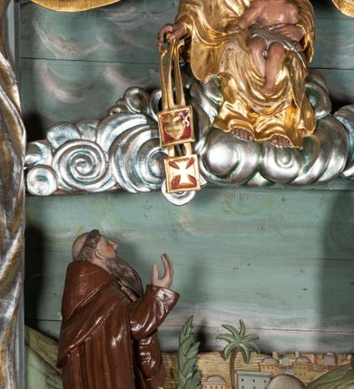Zdjęcie nr 5: Zwieńczenie ołtarza o fantazyjnym kształcie zbliżonym do stojącego prostokąta z płaskorzeźbioną sceną ofiarowania szkaplerza św. Szymonowi Stockowi. U góry zwieńczenia podpięta srebrzona kotara o złoconym brzegu, spływająca na jego boki. W górnej partii, po bokach para aniołków z wzniesionymi do góry rękami oraz dwie uskrzydlone główki anielskie. Na szczycie nastawy zatknięta jest promienista gloria złożona z wiązek promieni o nierównej długości z monogramem maryjnym otoczonym przez wieniec z obłoków. Pole zwieńczenia dwustrefowe, oddzielone pasem obłoków. W dolnej strefie, po lewej stronie rzeźba św. Szymona Stocka. Święty ukazany jest w pozycji klęczącej z prawego profilu z wniesionymi na wysokości piersi rękami i głową skierowaną ku Matce Bożej. Twarz podłużna z długim nosem i silnie zarumienionymi policzkami. Głowę okala długa i siwa broda oraz tonsura. Święty ubrany jest w brązowy habit i płaszcz karmelitański. Po prawej stronie obrazu płaskorzeźbione, zielone wzniesienia i duże drzewo. W tle malowany pejzaż miejski i jasnobłękitne niebo. W górnej strefie siedząca na obłokach Matka Boska z Dzieciątkiem. W prawej dłoni trzyma szkaplerze zdobione greckim krzyżykiem oraz gorejącym sercem, przebitym mieczem, a lewą ręką przytrzymuje siedzące na jej kolanach Dzieciątko Jezus. Maria ma podłużną twarz z niewielkimi ustami, długim nosem i jasnoniebieskimi oczami, okoloną ciemnobrązowymi włosami, opadającymi na plecy. Ubrana jest w złocone szaty: suknię i płaszcz, obficie drapowane. Na głowie ma założony srebrzony welon oraz pełną koronę, a wokół niej wieniec z 12 gwiazd. Polichromia struktury zwieńczenia w kolorze jasnoniebieskim, dekoracje ornamentalne oraz detale złocone i srebrzone. Polichromia w odsłoniętych partiach ciała figur naturalistyczna.

