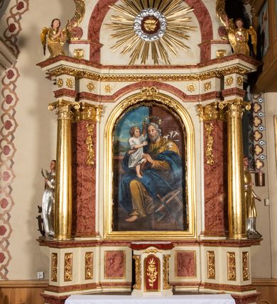 Zdjęcie nr 22: Nastawa ołtarzowa architektoniczna, jednoosiowa, jednokondygnacyjna ze zwieńczeniem. Ołtarz prostopadłościenny, drewniany z drewnianą mensą i z marmurowym portatylem. Antepedium w formie prostokąta ujętego profilowaną ramą. W polu marmoryzowana płycina o narożach zdobionych płaskorzeźbioną dekoracją roślinną złożoną z liści akantu oraz nałożonymi na siebie łacińskim krzyżem z kotwicą i gorejącym sercem oplecionym wicią z róż w polu. Tabernakulum w kształcie prostopadłościennej skrzynki na niskim cokole, flankowane dwoma pilastrami ustawionymi pod kątem, zdobionymi wicią z liści akantu; zamknięte od góry prostym belkowaniem, pośrodku falującym. Na drzwiczkach płycina w kształcie stojącego prostokąta, zamkniętego łukiem półkolistym, ujęta profilowaną ramą, w polu której umieszczone jest płaskorzeźbione gorejące serce osadzone na gałązce róży; tło czerwone.

Retabulum na zdwojonym cokole, flankowane ustawionymi pod kątem w jednej linii parami pilastrów, filarów oraz kolumn na przodzie. Podpory dźwigają wyłamane na osi belkowanie z ustawionymi na bokach rzeźbami aniołków. Trzony filarów są marmoryzowane; trzony kolumn delikatnie zwężające się ku górze, złocone, zdobione pionowymi pasami, co drugi wypełniony pnącą się ku górze rytą wicią z liści dębu lub z liści klonu; kapitele podpór kompozytowe.  Po bokach ołtarza na niskich cokołach ustawione pod kątem rzeźby św. Katarzyny Aleksandryjskiej po lewej i św. Wawrzyńca po prawej stronie. W polu, w głębokiej wnęce ujętej szeroką, profilowaną ramą umieszczony jest obraz św. Józefa z Dzieciątkiem. Wnęka od wewnątrz zdobiona płaskorzeźbioną winną latoroślą. Cokół dekorowany jest prostokątnymi, marmoryzowanymi płycinami; fryz złoconymi rozetami.

Zwieńczenie monumentalne, o fantazyjnym kształcie, po bokach ujęte wolutami zdobionymi liśćmi akantu, zamknięte od góry prostym gzymsem w formie nadwieszonego łuku z zatkniętym na szczycie łacińskim krzyżykiem. W polu, w półkolistej wnęce promienista gloria z dwoma gorejącymi sercami, z których jedno przebite jest mieczem, umieszczonym w otoku z obłoków.

Struktura w kolorze jasnobeżowym, płyciny, filary i detale marmoryzowana w kolorze różowym; ornamentyka i profilowania złocone i srebrzone.

