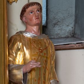 Zdjęcie nr 1: Rzeźba pełnoplastyczna ustawiona na niskim, szarym cokole o ściętych narożach. Święty Wawrzyniec ukazany jest frontalnie w delikatnym kontrapoście z lewą nogą ugiętą w kolanie. Prawą rękę ma ugiętą w łokciu i wysuniętą przed siebie, lewą wspiera na ruszcie opartym o cokół, głowę ma delikatnie uniesioną do góry. Twarz szeroka z małymi i wąskimi ustami, długim nosem, dużymi oczami ze wzrokiem skierowanym ku górze. Włosy krótkie, ciemnobrązowe z zaznaczoną tonsurą. Święty ubrany jest w długą, srebrzoną albę z długimi rękawami oraz złotą dalmatykę dekorowaną na pretekście pojedynczymi kwiatkami lilii. Polichromia naturalistyczna w odsłoniętych partiach ciała, strój złocony i srebrzony.