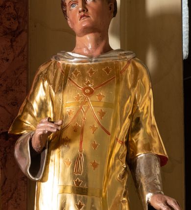 Zdjęcie nr 4: Rzeźba pełnoplastyczna ustawiona na niskim, szarym cokole o ściętych narożach. Święty Wawrzyniec ukazany jest frontalnie w delikatnym kontrapoście z lewą nogą ugiętą w kolanie. Prawą rękę ma ugiętą w łokciu i wysuniętą przed siebie, lewą wspiera na ruszcie opartym o cokół, głowę ma delikatnie uniesioną do góry. Twarz szeroka z małymi i wąskimi ustami, długim nosem, dużymi oczami ze wzrokiem skierowanym ku górze. Włosy krótkie, ciemnobrązowe z zaznaczoną tonsurą. Święty ubrany jest w długą, srebrzoną albę z długimi rękawami oraz złotą dalmatykę dekorowaną na pretekście pojedynczymi kwiatkami lilii. Polichromia naturalistyczna w odsłoniętych partiach ciała, strój złocony i srebrzony.