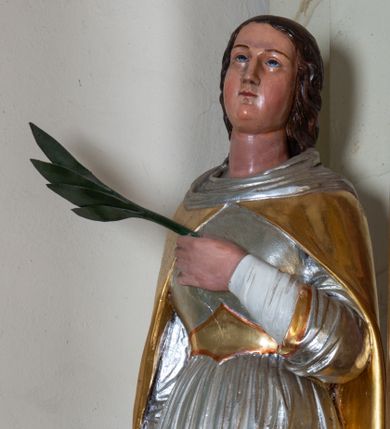 Zdjęcie nr 1: Rzeźba pełnoplastyczna ustawiona na szarym, niskim cokole o ściętych narożach. Święta Katarzyna Aleksandryjska ukazana jest frontalnie w delikatnym kontrapoście z lewą nogą ugiętą w kolanie. Prawą ręką wspiera się na dużym, złamanym kole, a w lewej dłoni złożonej na piersi trzyma palmę męczeństwa. Twarz szeroka z małymi i wąskimi ustami, długim nosem, małymi oczami ze wzrokiem skierowanym ku górze. Jej długie ciemnobrązowe włosy opadają na plecy i zasłaniają uszy. Święta ubrana jest w długą, srebrną suknię, przewiązaną w talii złotym pasem oraz zarzucony na plecy, złoty płaszcz, spięty pod szyją; na stopach ma założone czarne, pełne buty. Polichromia naturalistyczna w odsłoniętych partiach ciała, strój złocony i srebrzony.