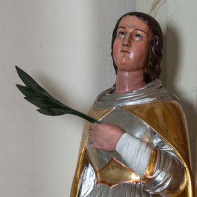 Zdjęcie nr 1: Rzeźba pełnoplastyczna ustawiona na szarym, niskim cokole o ściętych narożach. Święta Katarzyna Aleksandryjska ukazana jest frontalnie w delikatnym kontrapoście z lewą nogą ugiętą w kolanie. Prawą ręką wspiera się na dużym, złamanym kole, a w lewej dłoni złożonej na piersi trzyma palmę męczeństwa. Twarz szeroka z małymi i wąskimi ustami, długim nosem, małymi oczami ze wzrokiem skierowanym ku górze. Jej długie ciemnobrązowe włosy opadają na plecy i zasłaniają uszy. Święta ubrana jest w długą, srebrną suknię, przewiązaną w talii złotym pasem oraz zarzucony na plecy, złoty płaszcz, spięty pod szyją; na stopach ma założone czarne, pełne buty. Polichromia naturalistyczna w odsłoniętych partiach ciała, strój złocony i srebrzony.