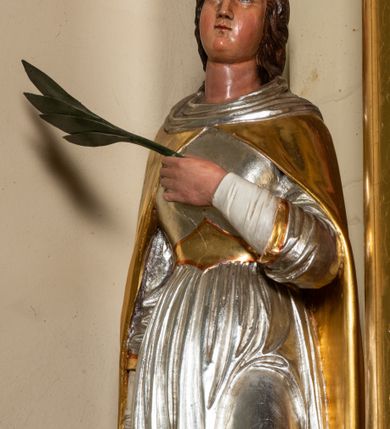 Zdjęcie nr 4: Rzeźba pełnoplastyczna ustawiona na szarym, niskim cokole o ściętych narożach. Święta Katarzyna Aleksandryjska ukazana jest frontalnie w delikatnym kontrapoście z lewą nogą ugiętą w kolanie. Prawą ręką wspiera się na dużym, złamanym kole, a w lewej dłoni złożonej na piersi trzyma palmę męczeństwa. Twarz szeroka z małymi i wąskimi ustami, długim nosem, małymi oczami ze wzrokiem skierowanym ku górze. Jej długie ciemnobrązowe włosy opadają na plecy i zasłaniają uszy. Święta ubrana jest w długą, srebrną suknię, przewiązaną w talii złotym pasem oraz zarzucony na plecy, złoty płaszcz, spięty pod szyją; na stopach ma założone czarne, pełne buty. Polichromia naturalistyczna w odsłoniętych partiach ciała, strój złocony i srebrzony.