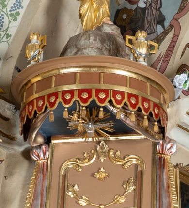 Zdjęcie nr 4: Ambona wisząca, przyścienna z wejściem od strony  kaplicy po północnej stronie prezbiterium. Kosz na planie półkolistym, na profilowanym cokole, z szyszką u spodu, flankowany po bokach dwoma spływami wolutowymi, na których ustawione są rzeźby siedzących aniołków. Na ściance kosza w profilowanej, złoconej ramie płaskorzeźba z przedstawieniem Połowu ryb. Zaplecek w kształcie prostokąta stojącego ujęty po bokach tkaniną przewiązaną u góry; w polu przedzielony złoconymi pasami ułożonymi na kształt litery H. W dolnej części zdobiony prostokątną płyciną ozdobioną płaskorzeźbioną rozetą, w górnej partii fantazyjną wicią roślinną. Baldachim  o wykroju półkolistym, w strefie belkowania zdobiony prostym fryzem przedzielonym pionowymi paskami na małe prostokątne płyciny, od dołu zdobiony lambrekinem, z dwoma wazonami płomienistymi z przodu oraz rzeźbą Mojżesza na skale w zwieńczeniu. U spodu baldachimu Gołębica Ducha Świętego w promienistej glorii złożonej z wiązek promieni o nierównej długości.
Płaskorzeźba na ściance frontowej kosza z przedstawieniem Połowu ryb. Kompozycja wielopostaciowa z ukazaniem po prawej stronie Jezusa nauczającego w łodzi oraz trzech uczniów wyciągających sieć pełną ryb. Po lewej stronie na brzegu siedząca postać jednego z uczniów. Uczniowie ubrani w długie suknie z długimi rękawami w kolorach: czerwonym, niebieskim i zielonym. Jezus ubrany jest w długą, złotą suknię z długim rękawem i złoty płaszcz. Krajobraz ukazany sumarycznie z wyraźnie zaznaczonymi falami morskimi. Tło złocone, ze złoconymi obłokami.
Aniołki siedzące na wolutach ukazane frontalnie, z  rozpostartymi szeroko skrzydłami. Aniołek po lewej stronie z rękami lekko ugiętymi w łokciach, z zamkniętą księgą w lewej dłoni. Aniołek po prawej stronie, delikatnie przechylony w lewo, z otwartą księgą w lewej dłoni i prawą ręką uniesioną nad księgą. Aniołki przepasane w pasie złotą chustą. Twarze szerokie, z pucułowatymi i delikatnie zarumienionymi policzkami, małe i wąskie usta, krótkie nosy. Włosy średniej długości, ciemnobrązowe, kędzierzawe.
W zwieńczeniu na wysokiej skale ustawiona jest rzeźba Mojżesza, ukazana frontalnie, z lewą nogą wspartą na skale, z tablicą przykazań w lewym ręku i laską w prawej. Mojżesz ubrany w długą, złotą suknię z długim rękawem. Twarz podłużna, okolona długą, ciemnobrązową brodą i krótkimi, ciemnobrązowymi włosami.
Struktura ambony polichromowana w kolorze jasnobrązowym, detale w kolorze ciemnobrązowym,  profile i ornamenty złocone oraz posrebrzone.