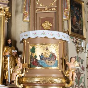 Zdjęcie nr 1: Ambona wisząca, przyścienna z wejściem od strony  kaplicy po północnej stronie prezbiterium. Kosz na planie półkolistym, na profilowanym cokole, z szyszką u spodu, flankowany po bokach dwoma spływami wolutowymi, na których ustawione są rzeźby siedzących aniołków. Na ściance kosza w profilowanej, złoconej ramie płaskorzeźba z przedstawieniem Połowu ryb. Zaplecek w kształcie prostokąta stojącego ujęty po bokach tkaniną przewiązaną u góry; w polu przedzielony złoconymi pasami ułożonymi na kształt litery H. W dolnej części zdobiony prostokątną płyciną ozdobioną płaskorzeźbioną rozetą, w górnej partii fantazyjną wicią roślinną. Baldachim  o wykroju półkolistym, w strefie belkowania zdobiony prostym fryzem przedzielonym pionowymi paskami na małe prostokątne płyciny, od dołu zdobiony lambrekinem, z dwoma wazonami płomienistymi z przodu oraz rzeźbą Mojżesza na skale w zwieńczeniu. U spodu baldachimu Gołębica Ducha Świętego w promienistej glorii złożonej z wiązek promieni o nierównej długości.
Płaskorzeźba na ściance frontowej kosza z przedstawieniem Połowu ryb. Kompozycja wielopostaciowa z ukazaniem po prawej stronie Jezusa nauczającego w łodzi oraz trzech uczniów wyciągających sieć pełną ryb. Po lewej stronie na brzegu siedząca postać jednego z uczniów. Uczniowie ubrani w długie suknie z długimi rękawami w kolorach: czerwonym, niebieskim i zielonym. Jezus ubrany jest w długą, złotą suknię z długim rękawem i złoty płaszcz. Krajobraz ukazany sumarycznie z wyraźnie zaznaczonymi falami morskimi. Tło złocone, ze złoconymi obłokami.
Aniołki siedzące na wolutach ukazane frontalnie, z  rozpostartymi szeroko skrzydłami. Aniołek po lewej stronie z rękami lekko ugiętymi w łokciach, z zamkniętą księgą w lewej dłoni. Aniołek po prawej stronie, delikatnie przechylony w lewo, z otwartą księgą w lewej dłoni i prawą ręką uniesioną nad księgą. Aniołki przepasane w pasie złotą chustą. Twarze szerokie, z pucułowatymi i delikatnie zarumienionymi policzkami, małe i wąskie usta, krótkie nosy. Włosy średniej długości, ciemnobrązowe, kędzierzawe.
W zwieńczeniu na wysokiej skale ustawiona jest rzeźba Mojżesza, ukazana frontalnie, z lewą nogą wspartą na skale, z tablicą przykazań w lewym ręku i laską w prawej. Mojżesz ubrany w długą, złotą suknię z długim rękawem. Twarz podłużna, okolona długą, ciemnobrązową brodą i krótkimi, ciemnobrązowymi włosami.
Struktura ambony polichromowana w kolorze jasnobrązowym, detale w kolorze ciemnobrązowym,  profile i ornamenty złocone oraz posrebrzone.