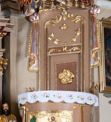 Zdjęcie nr 3: Ambona wisząca, przyścienna z wejściem od strony  kaplicy po północnej stronie prezbiterium. Kosz na planie półkolistym, na profilowanym cokole, z szyszką u spodu, flankowany po bokach dwoma spływami wolutowymi, na których ustawione są rzeźby siedzących aniołków. Na ściance kosza w profilowanej, złoconej ramie płaskorzeźba z przedstawieniem Połowu ryb. Zaplecek w kształcie prostokąta stojącego ujęty po bokach tkaniną przewiązaną u góry; w polu przedzielony złoconymi pasami ułożonymi na kształt litery H. W dolnej części zdobiony prostokątną płyciną ozdobioną płaskorzeźbioną rozetą, w górnej partii fantazyjną wicią roślinną. Baldachim  o wykroju półkolistym, w strefie belkowania zdobiony prostym fryzem przedzielonym pionowymi paskami na małe prostokątne płyciny, od dołu zdobiony lambrekinem, z dwoma wazonami płomienistymi z przodu oraz rzeźbą Mojżesza na skale w zwieńczeniu. U spodu baldachimu Gołębica Ducha Świętego w promienistej glorii złożonej z wiązek promieni o nierównej długości.
Płaskorzeźba na ściance frontowej kosza z przedstawieniem Połowu ryb. Kompozycja wielopostaciowa z ukazaniem po prawej stronie Jezusa nauczającego w łodzi oraz trzech uczniów wyciągających sieć pełną ryb. Po lewej stronie na brzegu siedząca postać jednego z uczniów. Uczniowie ubrani w długie suknie z długimi rękawami w kolorach: czerwonym, niebieskim i zielonym. Jezus ubrany jest w długą, złotą suknię z długim rękawem i złoty płaszcz. Krajobraz ukazany sumarycznie z wyraźnie zaznaczonymi falami morskimi. Tło złocone, ze złoconymi obłokami.
Aniołki siedzące na wolutach ukazane frontalnie, z  rozpostartymi szeroko skrzydłami. Aniołek po lewej stronie z rękami lekko ugiętymi w łokciach, z zamkniętą księgą w lewej dłoni. Aniołek po prawej stronie, delikatnie przechylony w lewo, z otwartą księgą w lewej dłoni i prawą ręką uniesioną nad księgą. Aniołki przepasane w pasie złotą chustą. Twarze szerokie, z pucułowatymi i delikatnie zarumienionymi policzkami, małe i wąskie usta, krótkie nosy. Włosy średniej długości, ciemnobrązowe, kędzierzawe.
W zwieńczeniu na wysokiej skale ustawiona jest rzeźba Mojżesza, ukazana frontalnie, z lewą nogą wspartą na skale, z tablicą przykazań w lewym ręku i laską w prawej. Mojżesz ubrany w długą, złotą suknię z długim rękawem. Twarz podłużna, okolona długą, ciemnobrązową brodą i krótkimi, ciemnobrązowymi włosami.
Struktura ambony polichromowana w kolorze jasnobrązowym, detale w kolorze ciemnobrązowym,  profile i ornamenty złocone oraz posrebrzone.