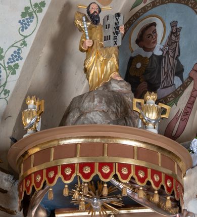 Zdjęcie nr 5: Ambona wisząca, przyścienna z wejściem od strony  kaplicy po północnej stronie prezbiterium. Kosz na planie półkolistym, na profilowanym cokole, z szyszką u spodu, flankowany po bokach dwoma spływami wolutowymi, na których ustawione są rzeźby siedzących aniołków. Na ściance kosza w profilowanej, złoconej ramie płaskorzeźba z przedstawieniem Połowu ryb. Zaplecek w kształcie prostokąta stojącego ujęty po bokach tkaniną przewiązaną u góry; w polu przedzielony złoconymi pasami ułożonymi na kształt litery H. W dolnej części zdobiony prostokątną płyciną ozdobioną płaskorzeźbioną rozetą, w górnej partii fantazyjną wicią roślinną. Baldachim  o wykroju półkolistym, w strefie belkowania zdobiony prostym fryzem przedzielonym pionowymi paskami na małe prostokątne płyciny, od dołu zdobiony lambrekinem, z dwoma wazonami płomienistymi z przodu oraz rzeźbą Mojżesza na skale w zwieńczeniu. U spodu baldachimu Gołębica Ducha Świętego w promienistej glorii złożonej z wiązek promieni o nierównej długości.
Płaskorzeźba na ściance frontowej kosza z przedstawieniem Połowu ryb. Kompozycja wielopostaciowa z ukazaniem po prawej stronie Jezusa nauczającego w łodzi oraz trzech uczniów wyciągających sieć pełną ryb. Po lewej stronie na brzegu siedząca postać jednego z uczniów. Uczniowie ubrani w długie suknie z długimi rękawami w kolorach: czerwonym, niebieskim i zielonym. Jezus ubrany jest w długą, złotą suknię z długim rękawem i złoty płaszcz. Krajobraz ukazany sumarycznie z wyraźnie zaznaczonymi falami morskimi. Tło złocone, ze złoconymi obłokami.
Aniołki siedzące na wolutach ukazane frontalnie, z  rozpostartymi szeroko skrzydłami. Aniołek po lewej stronie z rękami lekko ugiętymi w łokciach, z zamkniętą księgą w lewej dłoni. Aniołek po prawej stronie, delikatnie przechylony w lewo, z otwartą księgą w lewej dłoni i prawą ręką uniesioną nad księgą. Aniołki przepasane w pasie złotą chustą. Twarze szerokie, z pucułowatymi i delikatnie zarumienionymi policzkami, małe i wąskie usta, krótkie nosy. Włosy średniej długości, ciemnobrązowe, kędzierzawe.
W zwieńczeniu na wysokiej skale ustawiona jest rzeźba Mojżesza, ukazana frontalnie, z lewą nogą wspartą na skale, z tablicą przykazań w lewym ręku i laską w prawej. Mojżesz ubrany w długą, złotą suknię z długim rękawem. Twarz podłużna, okolona długą, ciemnobrązową brodą i krótkimi, ciemnobrązowymi włosami.
Struktura ambony polichromowana w kolorze jasnobrązowym, detale w kolorze ciemnobrązowym,  profile i ornamenty złocone oraz posrebrzone.