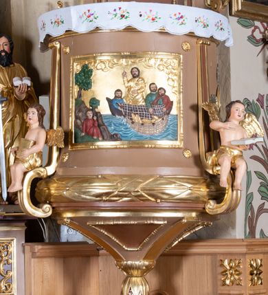 Zdjęcie nr 6: Ambona wisząca, przyścienna z wejściem od strony  kaplicy po północnej stronie prezbiterium. Kosz na planie półkolistym, na profilowanym cokole, z szyszką u spodu, flankowany po bokach dwoma spływami wolutowymi, na których ustawione są rzeźby siedzących aniołków. Na ściance kosza w profilowanej, złoconej ramie płaskorzeźba z przedstawieniem Połowu ryb. Zaplecek w kształcie prostokąta stojącego ujęty po bokach tkaniną przewiązaną u góry; w polu przedzielony złoconymi pasami ułożonymi na kształt litery H. W dolnej części zdobiony prostokątną płyciną ozdobioną płaskorzeźbioną rozetą, w górnej partii fantazyjną wicią roślinną. Baldachim  o wykroju półkolistym, w strefie belkowania zdobiony prostym fryzem przedzielonym pionowymi paskami na małe prostokątne płyciny, od dołu zdobiony lambrekinem, z dwoma wazonami płomienistymi z przodu oraz rzeźbą Mojżesza na skale w zwieńczeniu. U spodu baldachimu Gołębica Ducha Świętego w promienistej glorii złożonej z wiązek promieni o nierównej długości.
Płaskorzeźba na ściance frontowej kosza z przedstawieniem Połowu ryb. Kompozycja wielopostaciowa z ukazaniem po prawej stronie Jezusa nauczającego w łodzi oraz trzech uczniów wyciągających sieć pełną ryb. Po lewej stronie na brzegu siedząca postać jednego z uczniów. Uczniowie ubrani w długie suknie z długimi rękawami w kolorach: czerwonym, niebieskim i zielonym. Jezus ubrany jest w długą, złotą suknię z długim rękawem i złoty płaszcz. Krajobraz ukazany sumarycznie z wyraźnie zaznaczonymi falami morskimi. Tło złocone, ze złoconymi obłokami.
Aniołki siedzące na wolutach ukazane frontalnie, z  rozpostartymi szeroko skrzydłami. Aniołek po lewej stronie z rękami lekko ugiętymi w łokciach, z zamkniętą księgą w lewej dłoni. Aniołek po prawej stronie, delikatnie przechylony w lewo, z otwartą księgą w lewej dłoni i prawą ręką uniesioną nad księgą. Aniołki przepasane w pasie złotą chustą. Twarze szerokie, z pucułowatymi i delikatnie zarumienionymi policzkami, małe i wąskie usta, krótkie nosy. Włosy średniej długości, ciemnobrązowe, kędzierzawe.
W zwieńczeniu na wysokiej skale ustawiona jest rzeźba Mojżesza, ukazana frontalnie, z lewą nogą wspartą na skale, z tablicą przykazań w lewym ręku i laską w prawej. Mojżesz ubrany w długą, złotą suknię z długim rękawem. Twarz podłużna, okolona długą, ciemnobrązową brodą i krótkimi, ciemnobrązowymi włosami.
Struktura ambony polichromowana w kolorze jasnobrązowym, detale w kolorze ciemnobrązowym,  profile i ornamenty złocone oraz posrebrzone.