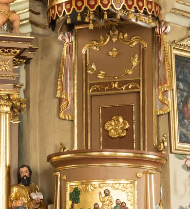 Zdjęcie nr 2: Ambona wisząca, przyścienna z wejściem od strony  kaplicy po północnej stronie prezbiterium. Kosz na planie półkolistym, na profilowanym cokole, z szyszką u spodu, flankowany po bokach dwoma spływami wolutowymi, na których ustawione są rzeźby siedzących aniołków. Na ściance kosza w profilowanej, złoconej ramie płaskorzeźba z przedstawieniem Połowu ryb. Zaplecek w kształcie prostokąta stojącego ujęty po bokach tkaniną przewiązaną u góry; w polu przedzielony złoconymi pasami ułożonymi na kształt litery H. W dolnej części zdobiony prostokątną płyciną ozdobioną płaskorzeźbioną rozetą, w górnej partii fantazyjną wicią roślinną. Baldachim  o wykroju półkolistym, w strefie belkowania zdobiony prostym fryzem przedzielonym pionowymi paskami na małe prostokątne płyciny, od dołu zdobiony lambrekinem, z dwoma wazonami płomienistymi z przodu oraz rzeźbą Mojżesza na skale w zwieńczeniu. U spodu baldachimu Gołębica Ducha Świętego w promienistej glorii złożonej z wiązek promieni o nierównej długości.
Płaskorzeźba na ściance frontowej kosza z przedstawieniem Połowu ryb. Kompozycja wielopostaciowa z ukazaniem po prawej stronie Jezusa nauczającego w łodzi oraz trzech uczniów wyciągających sieć pełną ryb. Po lewej stronie na brzegu siedząca postać jednego z uczniów. Uczniowie ubrani w długie suknie z długimi rękawami w kolorach: czerwonym, niebieskim i zielonym. Jezus ubrany jest w długą, złotą suknię z długim rękawem i złoty płaszcz. Krajobraz ukazany sumarycznie z wyraźnie zaznaczonymi falami morskimi. Tło złocone, ze złoconymi obłokami.
Aniołki siedzące na wolutach ukazane frontalnie, z  rozpostartymi szeroko skrzydłami. Aniołek po lewej stronie z rękami lekko ugiętymi w łokciach, z zamkniętą księgą w lewej dłoni. Aniołek po prawej stronie, delikatnie przechylony w lewo, z otwartą księgą w lewej dłoni i prawą ręką uniesioną nad księgą. Aniołki przepasane w pasie złotą chustą. Twarze szerokie, z pucułowatymi i delikatnie zarumienionymi policzkami, małe i wąskie usta, krótkie nosy. Włosy średniej długości, ciemnobrązowe, kędzierzawe.
W zwieńczeniu na wysokiej skale ustawiona jest rzeźba Mojżesza, ukazana frontalnie, z lewą nogą wspartą na skale, z tablicą przykazań w lewym ręku i laską w prawej. Mojżesz ubrany w długą, złotą suknię z długim rękawem. Twarz podłużna, okolona długą, ciemnobrązową brodą i krótkimi, ciemnobrązowymi włosami.
Struktura ambony polichromowana w kolorze jasnobrązowym, detale w kolorze ciemnobrązowym,  profile i ornamenty złocone oraz posrebrzone.