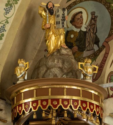 Zdjęcie nr 10: Ambona wisząca, przyścienna z wejściem od strony  kaplicy po północnej stronie prezbiterium. Kosz na planie półkolistym, na profilowanym cokole, z szyszką u spodu, flankowany po bokach dwoma spływami wolutowymi, na których ustawione są rzeźby siedzących aniołków. Na ściance kosza w profilowanej, złoconej ramie płaskorzeźba z przedstawieniem Połowu ryb. Zaplecek w kształcie prostokąta stojącego ujęty po bokach tkaniną przewiązaną u góry; w polu przedzielony złoconymi pasami ułożonymi na kształt litery H. W dolnej części zdobiony prostokątną płyciną ozdobioną płaskorzeźbioną rozetą, w górnej partii fantazyjną wicią roślinną. Baldachim  o wykroju półkolistym, w strefie belkowania zdobiony prostym fryzem przedzielonym pionowymi paskami na małe prostokątne płyciny, od dołu zdobiony lambrekinem, z dwoma wazonami płomienistymi z przodu oraz rzeźbą Mojżesza na skale w zwieńczeniu. U spodu baldachimu Gołębica Ducha Świętego w promienistej glorii złożonej z wiązek promieni o nierównej długości.
Płaskorzeźba na ściance frontowej kosza z przedstawieniem Połowu ryb. Kompozycja wielopostaciowa z ukazaniem po prawej stronie Jezusa nauczającego w łodzi oraz trzech uczniów wyciągających sieć pełną ryb. Po lewej stronie na brzegu siedząca postać jednego z uczniów. Uczniowie ubrani w długie suknie z długimi rękawami w kolorach: czerwonym, niebieskim i zielonym. Jezus ubrany jest w długą, złotą suknię z długim rękawem i złoty płaszcz. Krajobraz ukazany sumarycznie z wyraźnie zaznaczonymi falami morskimi. Tło złocone, ze złoconymi obłokami.
Aniołki siedzące na wolutach ukazane frontalnie, z  rozpostartymi szeroko skrzydłami. Aniołek po lewej stronie z rękami lekko ugiętymi w łokciach, z zamkniętą księgą w lewej dłoni. Aniołek po prawej stronie, delikatnie przechylony w lewo, z otwartą księgą w lewej dłoni i prawą ręką uniesioną nad księgą. Aniołki przepasane w pasie złotą chustą. Twarze szerokie, z pucułowatymi i delikatnie zarumienionymi policzkami, małe i wąskie usta, krótkie nosy. Włosy średniej długości, ciemnobrązowe, kędzierzawe.
W zwieńczeniu na wysokiej skale ustawiona jest rzeźba Mojżesza, ukazana frontalnie, z lewą nogą wspartą na skale, z tablicą przykazań w lewym ręku i laską w prawej. Mojżesz ubrany w długą, złotą suknię z długim rękawem. Twarz podłużna, okolona długą, ciemnobrązową brodą i krótkimi, ciemnobrązowymi włosami.
Struktura ambony polichromowana w kolorze jasnobrązowym, detale w kolorze ciemnobrązowym,  profile i ornamenty złocone oraz posrebrzone.