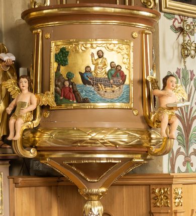 Zdjęcie nr 7: Ambona wisząca, przyścienna z wejściem od strony  kaplicy po północnej stronie prezbiterium. Kosz na planie półkolistym, na profilowanym cokole, z szyszką u spodu, flankowany po bokach dwoma spływami wolutowymi, na których ustawione są rzeźby siedzących aniołków. Na ściance kosza w profilowanej, złoconej ramie płaskorzeźba z przedstawieniem Połowu ryb. Zaplecek w kształcie prostokąta stojącego ujęty po bokach tkaniną przewiązaną u góry; w polu przedzielony złoconymi pasami ułożonymi na kształt litery H. W dolnej części zdobiony prostokątną płyciną ozdobioną płaskorzeźbioną rozetą, w górnej partii fantazyjną wicią roślinną. Baldachim  o wykroju półkolistym, w strefie belkowania zdobiony prostym fryzem przedzielonym pionowymi paskami na małe prostokątne płyciny, od dołu zdobiony lambrekinem, z dwoma wazonami płomienistymi z przodu oraz rzeźbą Mojżesza na skale w zwieńczeniu. U spodu baldachimu Gołębica Ducha Świętego w promienistej glorii złożonej z wiązek promieni o nierównej długości.
Płaskorzeźba na ściance frontowej kosza z przedstawieniem Połowu ryb. Kompozycja wielopostaciowa z ukazaniem po prawej stronie Jezusa nauczającego w łodzi oraz trzech uczniów wyciągających sieć pełną ryb. Po lewej stronie na brzegu siedząca postać jednego z uczniów. Uczniowie ubrani w długie suknie z długimi rękawami w kolorach: czerwonym, niebieskim i zielonym. Jezus ubrany jest w długą, złotą suknię z długim rękawem i złoty płaszcz. Krajobraz ukazany sumarycznie z wyraźnie zaznaczonymi falami morskimi. Tło złocone, ze złoconymi obłokami.
Aniołki siedzące na wolutach ukazane frontalnie, z  rozpostartymi szeroko skrzydłami. Aniołek po lewej stronie z rękami lekko ugiętymi w łokciach, z zamkniętą księgą w lewej dłoni. Aniołek po prawej stronie, delikatnie przechylony w lewo, z otwartą księgą w lewej dłoni i prawą ręką uniesioną nad księgą. Aniołki przepasane w pasie złotą chustą. Twarze szerokie, z pucułowatymi i delikatnie zarumienionymi policzkami, małe i wąskie usta, krótkie nosy. Włosy średniej długości, ciemnobrązowe, kędzierzawe.
W zwieńczeniu na wysokiej skale ustawiona jest rzeźba Mojżesza, ukazana frontalnie, z lewą nogą wspartą na skale, z tablicą przykazań w lewym ręku i laską w prawej. Mojżesz ubrany w długą, złotą suknię z długim rękawem. Twarz podłużna, okolona długą, ciemnobrązową brodą i krótkimi, ciemnobrązowymi włosami.
Struktura ambony polichromowana w kolorze jasnobrązowym, detale w kolorze ciemnobrązowym,  profile i ornamenty złocone oraz posrebrzone.