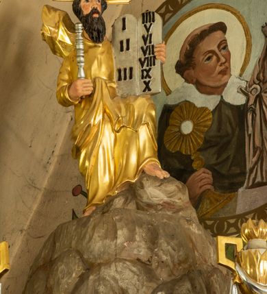 Zdjęcie nr 11: Ambona wisząca, przyścienna z wejściem od strony  kaplicy po północnej stronie prezbiterium. Kosz na planie półkolistym, na profilowanym cokole, z szyszką u spodu, flankowany po bokach dwoma spływami wolutowymi, na których ustawione są rzeźby siedzących aniołków. Na ściance kosza w profilowanej, złoconej ramie płaskorzeźba z przedstawieniem Połowu ryb. Zaplecek w kształcie prostokąta stojącego ujęty po bokach tkaniną przewiązaną u góry; w polu przedzielony złoconymi pasami ułożonymi na kształt litery H. W dolnej części zdobiony prostokątną płyciną ozdobioną płaskorzeźbioną rozetą, w górnej partii fantazyjną wicią roślinną. Baldachim  o wykroju półkolistym, w strefie belkowania zdobiony prostym fryzem przedzielonym pionowymi paskami na małe prostokątne płyciny, od dołu zdobiony lambrekinem, z dwoma wazonami płomienistymi z przodu oraz rzeźbą Mojżesza na skale w zwieńczeniu. U spodu baldachimu Gołębica Ducha Świętego w promienistej glorii złożonej z wiązek promieni o nierównej długości.
Płaskorzeźba na ściance frontowej kosza z przedstawieniem Połowu ryb. Kompozycja wielopostaciowa z ukazaniem po prawej stronie Jezusa nauczającego w łodzi oraz trzech uczniów wyciągających sieć pełną ryb. Po lewej stronie na brzegu siedząca postać jednego z uczniów. Uczniowie ubrani w długie suknie z długimi rękawami w kolorach: czerwonym, niebieskim i zielonym. Jezus ubrany jest w długą, złotą suknię z długim rękawem i złoty płaszcz. Krajobraz ukazany sumarycznie z wyraźnie zaznaczonymi falami morskimi. Tło złocone, ze złoconymi obłokami.
Aniołki siedzące na wolutach ukazane frontalnie, z  rozpostartymi szeroko skrzydłami. Aniołek po lewej stronie z rękami lekko ugiętymi w łokciach, z zamkniętą księgą w lewej dłoni. Aniołek po prawej stronie, delikatnie przechylony w lewo, z otwartą księgą w lewej dłoni i prawą ręką uniesioną nad księgą. Aniołki przepasane w pasie złotą chustą. Twarze szerokie, z pucułowatymi i delikatnie zarumienionymi policzkami, małe i wąskie usta, krótkie nosy. Włosy średniej długości, ciemnobrązowe, kędzierzawe.
W zwieńczeniu na wysokiej skale ustawiona jest rzeźba Mojżesza, ukazana frontalnie, z lewą nogą wspartą na skale, z tablicą przykazań w lewym ręku i laską w prawej. Mojżesz ubrany w długą, złotą suknię z długim rękawem. Twarz podłużna, okolona długą, ciemnobrązową brodą i krótkimi, ciemnobrązowymi włosami.
Struktura ambony polichromowana w kolorze jasnobrązowym, detale w kolorze ciemnobrązowym,  profile i ornamenty złocone oraz posrebrzone.