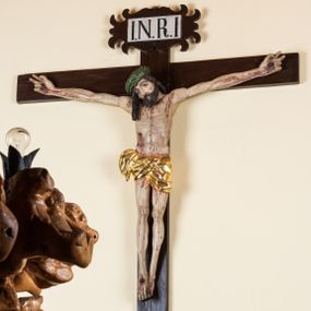 Zdjęcie nr 1: Rzeźba pełnoplastyczna przedstawiająca Chrystusa zawieszonego na krzyżu na wyprostowanych ramionach, figura w typie Cristo vivo. Głowa  Chrystusa delikatnie przechylona na prawe ramię; nogi wyprostowane w kolanach, stopy przebite jednym gwoździem w układzie prawa na lewą. Twarz Zbawiciela szeroka o wyraźnych rysach, długim i wąskim nosie oraz krótkiej brodzie, zawiniętej na końcu w dwa krótkie i silnie skręcone pukle. Włosy długie, opadające na plecy z jednym długim puklem spływającym sztywno na prawe ramię. Na głowie Chrystus ma założoną ciasno splecioną, zieloną koronę cierniową, zawiązaną opaską z gałązek. Sylwetka wychudzona, ułożona w nienaturalnej pozie z dużymi dłońmi i stopami. Perizonium złocone, ciasno oplatające biodra, przewiązane pośrodku z jednym końcem wystającym z prawej strony. Nad głową Chrystusa znajduje się titulus w formie białej tabliczki namalowanej na wolutowo wyciętym kartuszu z napisem w polu „I(ESUS) N(AZARENUS) R(EX) I[UDAEORUM]”. Polichromia naturalistyczna w odsłoniętych partiach ciała z zaznaczonymi śladami męki. Krzyż drewniany, prosty, malowany na kolor ciemnobrązowy.