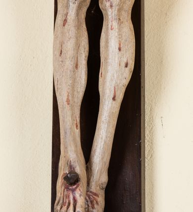 Zdjęcie nr 7: Rzeźba pełnoplastyczna przedstawiająca Chrystusa zawieszonego na krzyżu na wyprostowanych ramionach, figura w typie Cristo vivo. Głowa  Chrystusa delikatnie przechylona na prawe ramię; nogi wyprostowane w kolanach, stopy przebite jednym gwoździem w układzie prawa na lewą. Twarz Zbawiciela szeroka o wyraźnych rysach, długim i wąskim nosie oraz krótkiej brodzie, zawiniętej na końcu w dwa krótkie i silnie skręcone pukle. Włosy długie, opadające na plecy z jednym długim puklem spływającym sztywno na prawe ramię. Na głowie Chrystus ma założoną ciasno splecioną, zieloną koronę cierniową, zawiązaną opaską z gałązek. Sylwetka wychudzona, ułożona w nienaturalnej pozie z dużymi dłońmi i stopami. Perizonium złocone, ciasno oplatające biodra, przewiązane pośrodku z jednym końcem wystającym z prawej strony. Nad głową Chrystusa znajduje się titulus w formie białej tabliczki namalowanej na wolutowo wyciętym kartuszu z napisem w polu „I(ESUS) N(AZARENUS) R(EX) I[UDAEORUM]”. Polichromia naturalistyczna w odsłoniętych partiach ciała z zaznaczonymi śladami męki. Krzyż drewniany, prosty, malowany na kolor ciemnobrązowy.