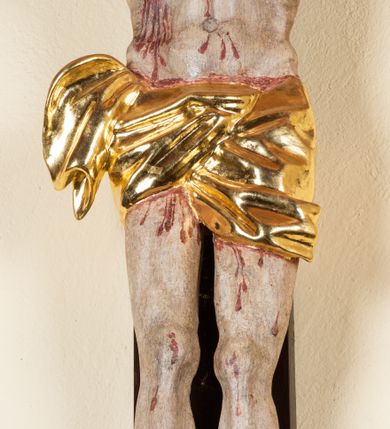 Zdjęcie nr 11: Rzeźba pełnoplastyczna przedstawiająca Chrystusa zawieszonego na krzyżu na wyprostowanych ramionach, figura w typie Cristo vivo. Głowa  Chrystusa delikatnie przechylona na prawe ramię; nogi wyprostowane w kolanach, stopy przebite jednym gwoździem w układzie prawa na lewą. Twarz Zbawiciela szeroka o wyraźnych rysach, długim i wąskim nosie oraz krótkiej brodzie, zawiniętej na końcu w dwa krótkie i silnie skręcone pukle. Włosy długie, opadające na plecy z jednym długim puklem spływającym sztywno na prawe ramię. Na głowie Chrystus ma założoną ciasno splecioną, zieloną koronę cierniową, zawiązaną opaską z gałązek. Sylwetka wychudzona, ułożona w nienaturalnej pozie z dużymi dłońmi i stopami. Perizonium złocone, ciasno oplatające biodra, przewiązane pośrodku z jednym końcem wystającym z prawej strony. Nad głową Chrystusa znajduje się titulus w formie białej tabliczki namalowanej na wolutowo wyciętym kartuszu z napisem w polu „I(ESUS) N(AZARENUS) R(EX) I[UDAEORUM]”. Polichromia naturalistyczna w odsłoniętych partiach ciała z zaznaczonymi śladami męki. Krzyż drewniany, prosty, malowany na kolor ciemnobrązowy.