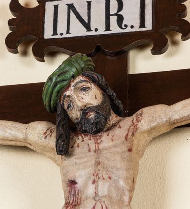 Zdjęcie nr 2: Rzeźba pełnoplastyczna przedstawiająca Chrystusa zawieszonego na krzyżu na wyprostowanych ramionach, figura w typie Cristo vivo. Głowa  Chrystusa delikatnie przechylona na prawe ramię; nogi wyprostowane w kolanach, stopy przebite jednym gwoździem w układzie prawa na lewą. Twarz Zbawiciela szeroka o wyraźnych rysach, długim i wąskim nosie oraz krótkiej brodzie, zawiniętej na końcu w dwa krótkie i silnie skręcone pukle. Włosy długie, opadające na plecy z jednym długim puklem spływającym sztywno na prawe ramię. Na głowie Chrystus ma założoną ciasno splecioną, zieloną koronę cierniową, zawiązaną opaską z gałązek. Sylwetka wychudzona, ułożona w nienaturalnej pozie z dużymi dłońmi i stopami. Perizonium złocone, ciasno oplatające biodra, przewiązane pośrodku z jednym końcem wystającym z prawej strony. Nad głową Chrystusa znajduje się titulus w formie białej tabliczki namalowanej na wolutowo wyciętym kartuszu z napisem w polu „I(ESUS) N(AZARENUS) R(EX) I[UDAEORUM]”. Polichromia naturalistyczna w odsłoniętych partiach ciała z zaznaczonymi śladami męki. Krzyż drewniany, prosty, malowany na kolor ciemnobrązowy.