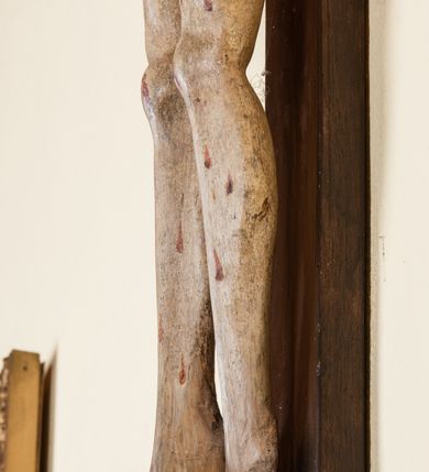 Zdjęcie nr 12: Rzeźba pełnoplastyczna przedstawiająca Chrystusa zawieszonego na krzyżu na wyprostowanych ramionach, figura w typie Cristo vivo. Głowa  Chrystusa delikatnie przechylona na prawe ramię; nogi wyprostowane w kolanach, stopy przebite jednym gwoździem w układzie prawa na lewą. Twarz Zbawiciela szeroka o wyraźnych rysach, długim i wąskim nosie oraz krótkiej brodzie, zawiniętej na końcu w dwa krótkie i silnie skręcone pukle. Włosy długie, opadające na plecy z jednym długim puklem spływającym sztywno na prawe ramię. Na głowie Chrystus ma założoną ciasno splecioną, zieloną koronę cierniową, zawiązaną opaską z gałązek. Sylwetka wychudzona, ułożona w nienaturalnej pozie z dużymi dłońmi i stopami. Perizonium złocone, ciasno oplatające biodra, przewiązane pośrodku z jednym końcem wystającym z prawej strony. Nad głową Chrystusa znajduje się titulus w formie białej tabliczki namalowanej na wolutowo wyciętym kartuszu z napisem w polu „I(ESUS) N(AZARENUS) R(EX) I[UDAEORUM]”. Polichromia naturalistyczna w odsłoniętych partiach ciała z zaznaczonymi śladami męki. Krzyż drewniany, prosty, malowany na kolor ciemnobrązowy.