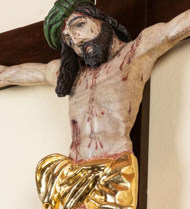 Zdjęcie nr 10: Rzeźba pełnoplastyczna przedstawiająca Chrystusa zawieszonego na krzyżu na wyprostowanych ramionach, figura w typie Cristo vivo. Głowa  Chrystusa delikatnie przechylona na prawe ramię; nogi wyprostowane w kolanach, stopy przebite jednym gwoździem w układzie prawa na lewą. Twarz Zbawiciela szeroka o wyraźnych rysach, długim i wąskim nosie oraz krótkiej brodzie, zawiniętej na końcu w dwa krótkie i silnie skręcone pukle. Włosy długie, opadające na plecy z jednym długim puklem spływającym sztywno na prawe ramię. Na głowie Chrystus ma założoną ciasno splecioną, zieloną koronę cierniową, zawiązaną opaską z gałązek. Sylwetka wychudzona, ułożona w nienaturalnej pozie z dużymi dłońmi i stopami. Perizonium złocone, ciasno oplatające biodra, przewiązane pośrodku z jednym końcem wystającym z prawej strony. Nad głową Chrystusa znajduje się titulus w formie białej tabliczki namalowanej na wolutowo wyciętym kartuszu z napisem w polu „I(ESUS) N(AZARENUS) R(EX) I[UDAEORUM]”. Polichromia naturalistyczna w odsłoniętych partiach ciała z zaznaczonymi śladami męki. Krzyż drewniany, prosty, malowany na kolor ciemnobrązowy.