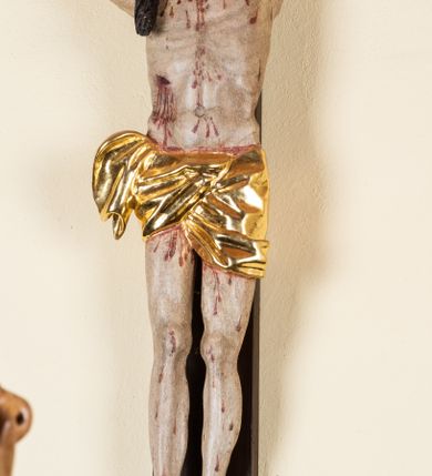Zdjęcie nr 9: Rzeźba pełnoplastyczna przedstawiająca Chrystusa zawieszonego na krzyżu na wyprostowanych ramionach, figura w typie Cristo vivo. Głowa  Chrystusa delikatnie przechylona na prawe ramię; nogi wyprostowane w kolanach, stopy przebite jednym gwoździem w układzie prawa na lewą. Twarz Zbawiciela szeroka o wyraźnych rysach, długim i wąskim nosie oraz krótkiej brodzie, zawiniętej na końcu w dwa krótkie i silnie skręcone pukle. Włosy długie, opadające na plecy z jednym długim puklem spływającym sztywno na prawe ramię. Na głowie Chrystus ma założoną ciasno splecioną, zieloną koronę cierniową, zawiązaną opaską z gałązek. Sylwetka wychudzona, ułożona w nienaturalnej pozie z dużymi dłońmi i stopami. Perizonium złocone, ciasno oplatające biodra, przewiązane pośrodku z jednym końcem wystającym z prawej strony. Nad głową Chrystusa znajduje się titulus w formie białej tabliczki namalowanej na wolutowo wyciętym kartuszu z napisem w polu „I(ESUS) N(AZARENUS) R(EX) I[UDAEORUM]”. Polichromia naturalistyczna w odsłoniętych partiach ciała z zaznaczonymi śladami męki. Krzyż drewniany, prosty, malowany na kolor ciemnobrązowy.