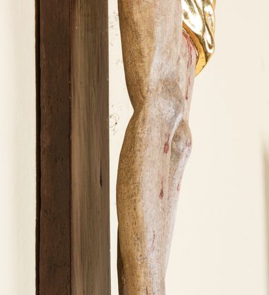 Zdjęcie nr 13: Rzeźba pełnoplastyczna przedstawiająca Chrystusa zawieszonego na krzyżu na wyprostowanych ramionach, figura w typie Cristo vivo. Głowa  Chrystusa delikatnie przechylona na prawe ramię; nogi wyprostowane w kolanach, stopy przebite jednym gwoździem w układzie prawa na lewą. Twarz Zbawiciela szeroka o wyraźnych rysach, długim i wąskim nosie oraz krótkiej brodzie, zawiniętej na końcu w dwa krótkie i silnie skręcone pukle. Włosy długie, opadające na plecy z jednym długim puklem spływającym sztywno na prawe ramię. Na głowie Chrystus ma założoną ciasno splecioną, zieloną koronę cierniową, zawiązaną opaską z gałązek. Sylwetka wychudzona, ułożona w nienaturalnej pozie z dużymi dłońmi i stopami. Perizonium złocone, ciasno oplatające biodra, przewiązane pośrodku z jednym końcem wystającym z prawej strony. Nad głową Chrystusa znajduje się titulus w formie białej tabliczki namalowanej na wolutowo wyciętym kartuszu z napisem w polu „I(ESUS) N(AZARENUS) R(EX) I[UDAEORUM]”. Polichromia naturalistyczna w odsłoniętych partiach ciała z zaznaczonymi śladami męki. Krzyż drewniany, prosty, malowany na kolor ciemnobrązowy.