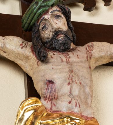 Zdjęcie nr 5: Rzeźba pełnoplastyczna przedstawiająca Chrystusa zawieszonego na krzyżu na wyprostowanych ramionach, figura w typie Cristo vivo. Głowa  Chrystusa delikatnie przechylona na prawe ramię; nogi wyprostowane w kolanach, stopy przebite jednym gwoździem w układzie prawa na lewą. Twarz Zbawiciela szeroka o wyraźnych rysach, długim i wąskim nosie oraz krótkiej brodzie, zawiniętej na końcu w dwa krótkie i silnie skręcone pukle. Włosy długie, opadające na plecy z jednym długim puklem spływającym sztywno na prawe ramię. Na głowie Chrystus ma założoną ciasno splecioną, zieloną koronę cierniową, zawiązaną opaską z gałązek. Sylwetka wychudzona, ułożona w nienaturalnej pozie z dużymi dłońmi i stopami. Perizonium złocone, ciasno oplatające biodra, przewiązane pośrodku z jednym końcem wystającym z prawej strony. Nad głową Chrystusa znajduje się titulus w formie białej tabliczki namalowanej na wolutowo wyciętym kartuszu z napisem w polu „I(ESUS) N(AZARENUS) R(EX) I[UDAEORUM]”. Polichromia naturalistyczna w odsłoniętych partiach ciała z zaznaczonymi śladami męki. Krzyż drewniany, prosty, malowany na kolor ciemnobrązowy.
