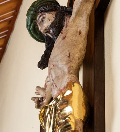 Zdjęcie nr 4: Rzeźba pełnoplastyczna przedstawiająca Chrystusa zawieszonego na krzyżu na wyprostowanych ramionach, figura w typie Cristo vivo. Głowa  Chrystusa delikatnie przechylona na prawe ramię; nogi wyprostowane w kolanach, stopy przebite jednym gwoździem w układzie prawa na lewą. Twarz Zbawiciela szeroka o wyraźnych rysach, długim i wąskim nosie oraz krótkiej brodzie, zawiniętej na końcu w dwa krótkie i silnie skręcone pukle. Włosy długie, opadające na plecy z jednym długim puklem spływającym sztywno na prawe ramię. Na głowie Chrystus ma założoną ciasno splecioną, zieloną koronę cierniową, zawiązaną opaską z gałązek. Sylwetka wychudzona, ułożona w nienaturalnej pozie z dużymi dłońmi i stopami. Perizonium złocone, ciasno oplatające biodra, przewiązane pośrodku z jednym końcem wystającym z prawej strony. Nad głową Chrystusa znajduje się titulus w formie białej tabliczki namalowanej na wolutowo wyciętym kartuszu z napisem w polu „I(ESUS) N(AZARENUS) R(EX) I[UDAEORUM]”. Polichromia naturalistyczna w odsłoniętych partiach ciała z zaznaczonymi śladami męki. Krzyż drewniany, prosty, malowany na kolor ciemnobrązowy.