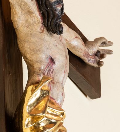 Zdjęcie nr 14: Rzeźba pełnoplastyczna przedstawiająca Chrystusa zawieszonego na krzyżu na wyprostowanych ramionach, figura w typie Cristo vivo. Głowa  Chrystusa delikatnie przechylona na prawe ramię; nogi wyprostowane w kolanach, stopy przebite jednym gwoździem w układzie prawa na lewą. Twarz Zbawiciela szeroka o wyraźnych rysach, długim i wąskim nosie oraz krótkiej brodzie, zawiniętej na końcu w dwa krótkie i silnie skręcone pukle. Włosy długie, opadające na plecy z jednym długim puklem spływającym sztywno na prawe ramię. Na głowie Chrystus ma założoną ciasno splecioną, zieloną koronę cierniową, zawiązaną opaską z gałązek. Sylwetka wychudzona, ułożona w nienaturalnej pozie z dużymi dłońmi i stopami. Perizonium złocone, ciasno oplatające biodra, przewiązane pośrodku z jednym końcem wystającym z prawej strony. Nad głową Chrystusa znajduje się titulus w formie białej tabliczki namalowanej na wolutowo wyciętym kartuszu z napisem w polu „I(ESUS) N(AZARENUS) R(EX) I[UDAEORUM]”. Polichromia naturalistyczna w odsłoniętych partiach ciała z zaznaczonymi śladami męki. Krzyż drewniany, prosty, malowany na kolor ciemnobrązowy.
