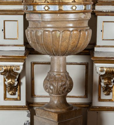 Zdjęcie nr 1: Chrzcielnica na podstawie na planie koła, ustawiona na wysokim cokole w formie sześcianu i na niskiej prostopadłościennej kostce. Trzon w dolnej części wklęsły; nodus w formie wydłużonej kuli dekorowanej liśćmi akantu. Czara półkolista, puklowana, o wklęsło-wypukłym brzegu. Wewnątrz metalowa misa. Pokrywa w formie korony z drewnianą imitacją kameryzowania obręczy i 18 sytuowanymi naprzemiennie kwiatonami i pałkami oraz siedmiu kabłąkami, podtrzymującymi kulę z krzyżem. Pokrywa złocona i srebrzona, od wewnątrz malowana na czerwono.