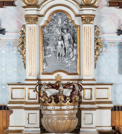 Zdjęcie nr 2: Chrzcielnica na podstawie na planie koła, ustawiona na wysokim cokole w formie sześcianu i na niskiej prostopadłościennej kostce. Trzon w dolnej części wklęsły; nodus w formie wydłużonej kuli dekorowanej liśćmi akantu. Czara półkolista, puklowana, o wklęsło-wypukłym brzegu. Wewnątrz metalowa misa. Pokrywa w formie korony z drewnianą imitacją kameryzowania obręczy i 18 sytuowanymi naprzemiennie kwiatonami i pałkami oraz siedmiu kabłąkami, podtrzymującymi kulę z krzyżem. Pokrywa złocona i srebrzona, od wewnątrz malowana na czerwono.