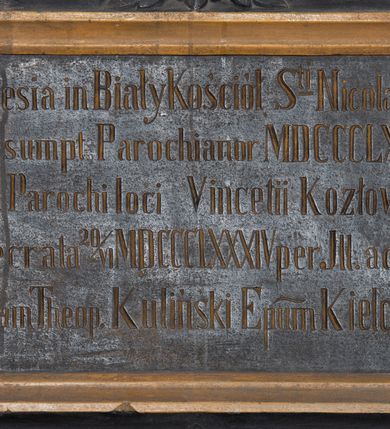 Zdjęcie nr 2: Tablica pamiątkowa w kształcie leżącego prostokąta, w formie kartusza, od dołu i od góry ujęta spływami wolutowymi. W polu płycina o uszowatych narożach z inskrypcją „Ec[c]lesia in Biały Kościół S(anc)ti Nicolai Epi(scopi) / aedif(icata) sumpt(u) Parochianor[um] MDCCCLXXVII / cura Parochi loci Vincentii Kozłowski / Consecrata 20 VI  MDCCCLXXXIV per Ill(ustrissimum) ac Rever(endissimum) / Thomam Theop(hilem) Kuliński Ep(iscop)um Kielcensem.” W zwieńczeniu dwie gałązki akantu. Całość malowana w kolorze czarnym, detale i profilowanie w kolorze jasnobrązowym.