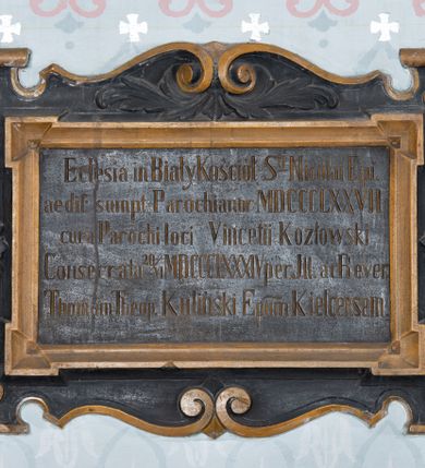 Zdjęcie nr 1: Tablica pamiątkowa w kształcie leżącego prostokąta, w formie kartusza, od dołu i od góry ujęta spływami wolutowymi. W polu płycina o uszowatych narożach z inskrypcją „Ec[c]lesia in Biały Kościół S(anc)ti Nicolai Epi(scopi) / aedif(icata) sumpt(u) Parochianor[um] MDCCCLXXVII / cura Parochi loci Vincentii Kozłowski / Consecrata 20 VI  MDCCCLXXXIV per Ill(ustrissimum) ac Rever(endissimum) / Thomam Theop(hilem) Kuliński Ep(iscop)um Kielcensem.” W zwieńczeniu dwie gałązki akantu. Całość malowana w kolorze czarnym, detale i profilowanie w kolorze jasnobrązowym.