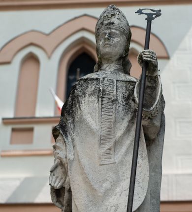 Zdjęcie nr 1: Rzeźba przedstawiająca św. Stanisława z Piotrowinem. Święty ukazany w postawie stojącej, frontalnie, w delikatnym kontrapoście z lewą nogą ugiętą w kolanie, z prawą ręką opuszczoną wzdłuż boku, w lewej trzyma pastorał. Twarz szeroka z drobnymi ustami, długim i wąskim nosem, niewielkimi oczami ze wzrokiem skierowanym na wprost. Święty ubrany jest w sutannę, rokietę zdobioną na brzegach koronką, ornat z pretekstą kolumnową, zdobioną poziomym żłobkowaniem, rękawiczki oraz  infułę z greckim krzyżykiem z przodu. U dołu, po lewej stronie kompozycji  Piotrowin, ukazany w półpostaci, nagi, wsparty na prawej ręce ugiętej w łokciu, z głową wzniesioną ku górze, z całunem zakrywającym włosy.