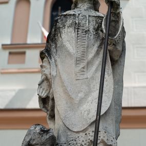 Zdjęcie nr 1: Rzeźba przedstawiająca św. Stanisława z Piotrowinem. Święty ukazany w postawie stojącej, frontalnie, w delikatnym kontrapoście z lewą nogą ugiętą w kolanie, z prawą ręką opuszczoną wzdłuż boku, w lewej trzyma pastorał. Twarz szeroka z drobnymi ustami, długim i wąskim nosem, niewielkimi oczami ze wzrokiem skierowanym na wprost. Święty ubrany jest w sutannę, rokietę zdobioną na brzegach koronką, ornat z pretekstą kolumnową, zdobioną poziomym żłobkowaniem, rękawiczki oraz  infułę z greckim krzyżykiem z przodu. U dołu, po lewej stronie kompozycji  Piotrowin, ukazany w półpostaci, nagi, wsparty na prawej ręce ugiętej w łokciu, z głową wzniesioną ku górze, z całunem zakrywającym włosy.