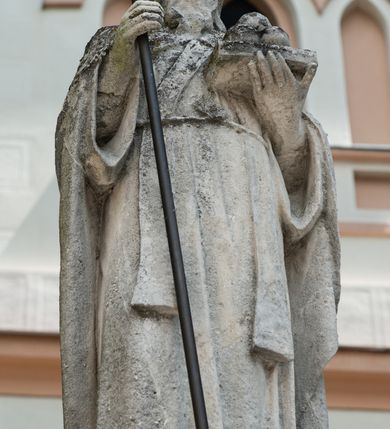 Zdjęcie nr 1: Rzeźba przedstawiająca św. Mikołaja. Święty ukazany jest w postawie stojącej, frontalnie, z krzyżem w prawej dłoni i otwartą księgą w lewej, na której ułożone są trzy kule. Twarz podłużna, okolona długą i bujną brodą, nos długi i wąski, oczy osadzone głęboko w oczodołach ze wzrokiem skierowanym na wprost; włosy długie i opadające na plecy. Święty ubrany jest w długą, przewiązaną sznurem albę, stułę, kapę zawiązaną pod szyją, infułę na głowie, zdobioną na przodzie krzyżykiem greckim, a na dłoniach ma założone rękawiczki. Krzyż na długim, metalowym drzewcu o ramionach zakończonych trójlistnie.